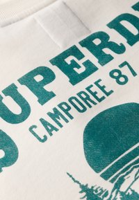Biały sweatshirt z tekstem w kolorze turkusowym, ekranowo drukowanym, z napisem "SUPERDRY CAMPOREE 87", zawierający graficzne elementy w postaci drzewa i okrągłego kształtu.