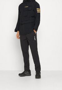 Veste imperméable noire avec poches avant et bandes dorées sur les manches, associée à un pantalon noir avec un logo sur le côté. Chaussures de sport noires.