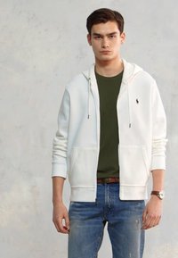 Jauns vīrietis, ģērbies baltā ar rāvējslēdzēju, olīvu zaļā T-kreklā, zilos džinsos un ar rokas pulksteni, stāv pretī vienkāršai gaiši krāsotai sienai.