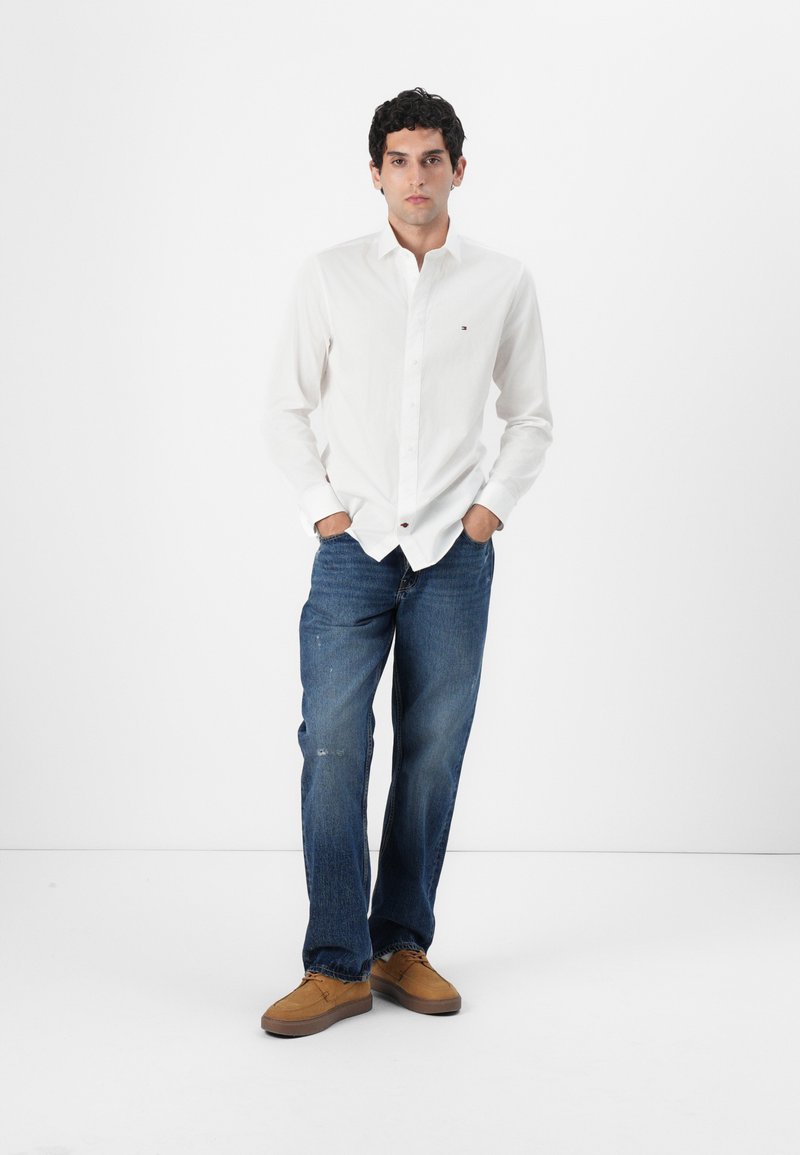Chemise blanche à boutons, jean bleu délavé et chaussures de couleur brun clair. La chemise arbore un petit logo ; le jean a une coupe décontractée et un léger effet usé.