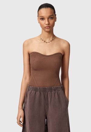 Jonge vrouw met naar achteren getrokken haar, draagt een strapless geribd bruin topje, losse bruine broek en een fijne ketting, staand met handen in de zakken.