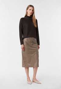 Frau in einem schwarzen Langarmpullover, einem taubfarbenen, knielangen Ledrrock mit Taschen und beigen spitz zulaufenden Slingback-Ballerinas, stehend vor weißem Hintergrund.