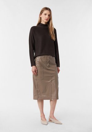 Frau in einem schwarzen Langarmpullover, einem taubfarbenen, knielangen Ledrrock mit Taschen und beigen spitz zulaufenden Slingback-Ballerinas, stehend vor weißem Hintergrund.