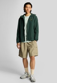 Chaqueta ligera verde con puños elásticos, camiseta de color azul claro debajo, pantalones cortos beige de carga y zapatillas deportivas grises.