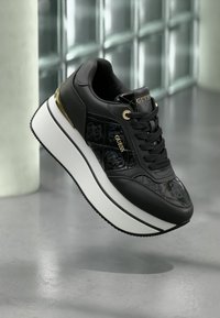 Sneakers Guess noirs et dorés avec une semelle blanche épaisse, des panneaux latéraux à motifs et des lacets noirs flottant au-dessus d'un sol gris.