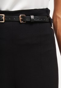 Zwarte broek met een gladde stofafwerking, een brede tailleband en een zwarte leren riem met goudkleurige hardware.