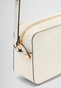 Sac bandoulière texturé blanc avec fermeture éclair dorée, bordure en cuir et logo gaufré. Présente un design slim et une bandoulière ajustable.