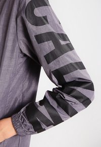 Šedá bunda s texturovaným povrchem, s velkým černým grafickým potiskem na levém rukávu a elastickými manžetami.