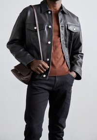 Svart läderjacka med tryckknappar, ljusbrun tröja, mörk denimjeans och en liten brun crossbody-väska. Slät textur och skräddarsydd passform.