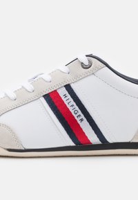 Weißer synthetischer Sneaker mit beigen Akzenten, versehen mit roten und marineblauen Streifen. Enthält Schnürsenkel und ein Metall-Logo-Emblem an der Ferse.