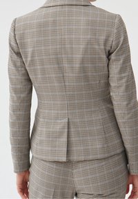 Ruskea ja beige ruudullinen bleiseri, jossa on pitkät hihat, yksittäinen halkio takana ja muotoillut olkapäät. Hienosti viimeistelty malli ja teksturoitu kangas.