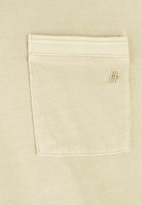 Beige shirtstof met een zachte textuur, voorzien van een gestikte patchzak en een subtiele geborduurde logo in een bijpassende kleur.