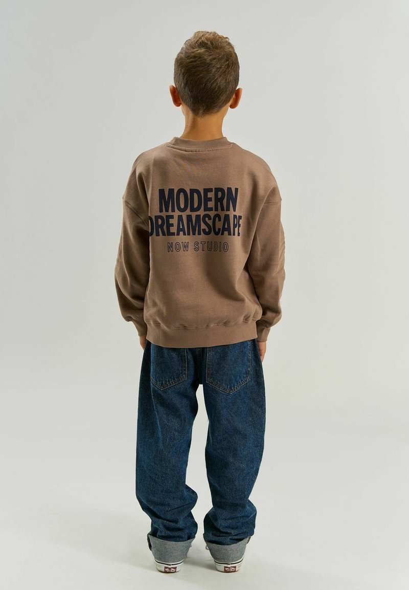 Enfant de dos portant un sweat-shirt marron avec le texte « MODERN DREAMSCAPE NOW STUDIO », un jean bleu et des baskets grises sur un fond clair.