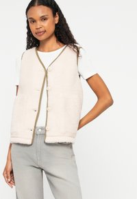 Gilet en polaire rose clair avec une bordure marron, doté d'une fermeture à boutons et de deux poches. Porté par-dessus une chemise blanche et associé à un jean gris.