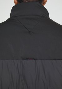 Vue de dos d'une personne portant une veste matelassée noire avec un col montant et une petite étiquette rouge, blanche et bleue sur le haut du dos.