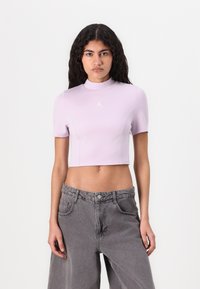 Lila rövid ujjú cropped top, magas nyakkivágással, a mellkason egy kis fehér logóval. Szürke, bő szárú farmerral párosítva.