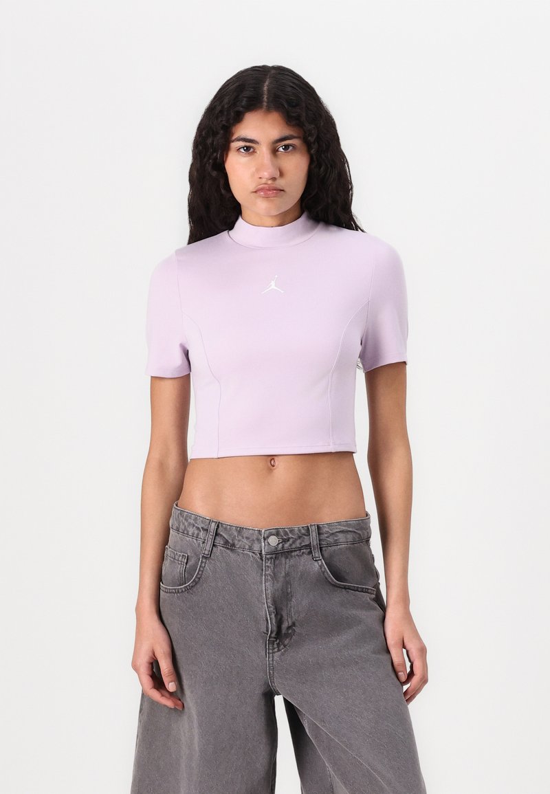 Lila rövid ujjú cropped top, magas nyakkivágással, a mellkason egy kis fehér logóval. Szürke, bő szárú farmerral párosítva.