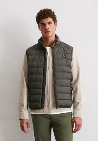 Olivgrüne Steppweste mit hohem Kragen, horizontalen Nähten, Reißverschluss vorne und einem kleinen Logopatch auf der Brust, getragen über einem hellbeigen Hemd.