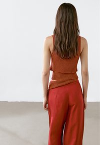 Hnědý pletený top s ribbed texturou, spárovaný s vysokými, splývavými červenými lněnými kalhotami, z pohledu zezadu. Jednoduchý design a pohodlný střih.