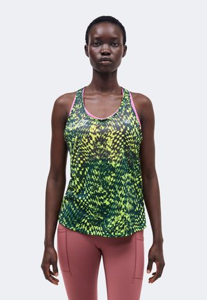 Kvinde med kort hår iført en grøn og gul mønstret ærmeløs sportstop og støvet lyserøde leggings, stående mod en ensfarvet baggrund.