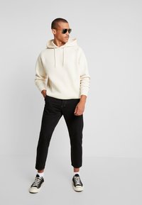 Felpa chiara con cappuccio e cordini, jeans neri slim-fit e sneakers bianco-neri. Modello con occhiali da sole in piedi contro uno sfondo neutro.