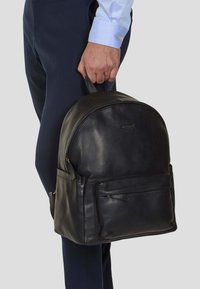 Svart läderryggsäck med rundad topp, ett handtag, framficka med dragkedja och två sidofickor. Slät textur och diskret design.