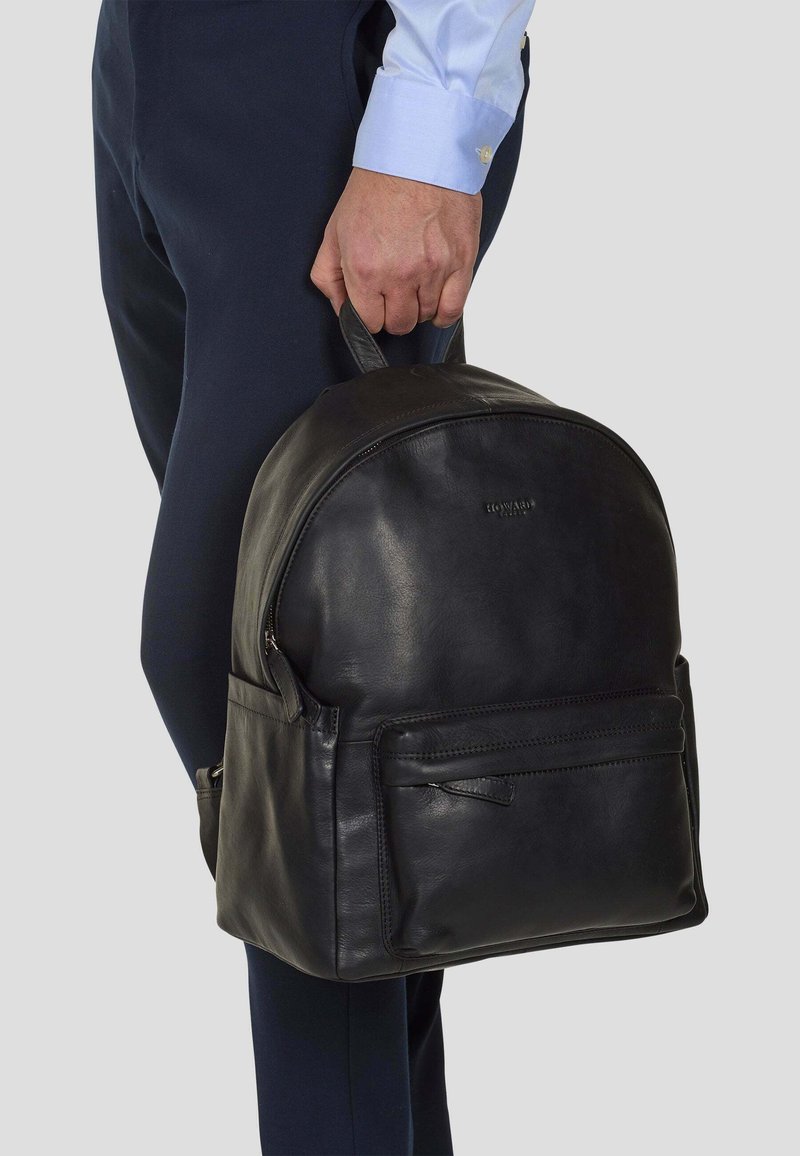 Svart läderryggsäck med rundad topp, ett handtag, framficka med dragkedja och två sidofickor. Slät textur och diskret design.