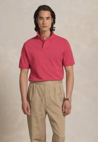 Νεαρός άνδρας με κόκκινο polo μπλουζάκι, μπεζ πλαϊνές παντελόνες και ρολόι χειρός, που στέκεται μπροστά από ένα ουδέτερο μπεζ φόντο.