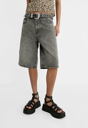Personne portant un short en jean gris au genou, ceinture noire cloutée, haut à imprimé léopard et sandales plateformes noires à brides.