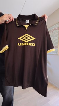 Hnedé polo tričko s žltými akcentmi, s vyšitým logom "UMBRO" a textúrou vdiamantovom vzore. Krátke rukávy, štandardný golier.