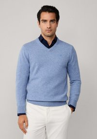 Světle modrý svetr s v-výstřihem vyrobený z měkkého materiálu s žebrovanými manžetami a lemem, nošený přes tmavou košili; decentní logo detail na prsou.