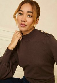 Brauner gestrickter Pullover mit hohem Kragen, gewellten Säumen und puffigen Ärmeln, der dekorative Knöpfe an der Schulter aufweist.