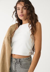 Vitt crop-top med hög halsringning, kombinerat med högmidjade, urtvättade grå denimjeans. Modellen håller en beige mockajacka.
