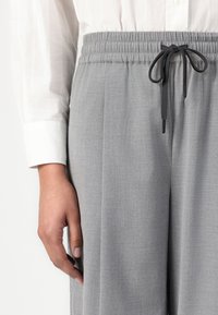 Prim-plan cu o persoană purtând pantaloni gri deschis cu talie elastică și șnur negru și o cămașă albă cu mâneci lungi, mâna relaxată pe lângă corp.