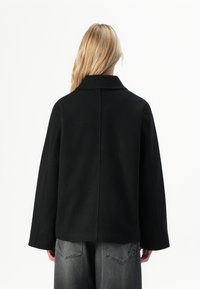 Person med langt blondt hår iført en svart jakke og falmede svarte jeans, vendende bort fra en ensfarget hvit bakgrunn.