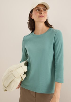 T-shirt à manches longues turquoise avec texture côtelée, col rond, associé à un sac beige clair. Design simple et moderne.