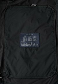 Zwarte opvouwbare jas met ritssluiting aan de binnenkant; voorzien van een label met diagrammen die vouw- en pakinstructies weergeven.