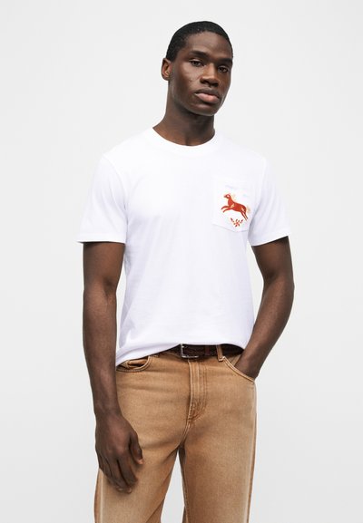 Νέος άνδρας φορούντας λευκό μπλουζάκι με κόκκινο σχέδιο αλόγου στην τσέπη στο στήθος και ανοιχτό καφέ τζιν με υφαντή ζώνη.