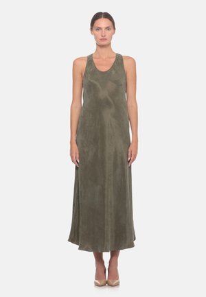 Donna che indossa un abito senza maniche verde oliva lungo fino a terra e tacchi alti beige, in piedi davanti a uno sfondo bianco uniforme.