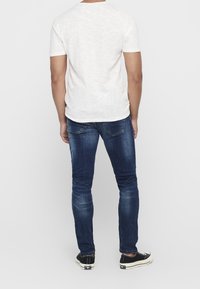 Vit t-shirt med kort ärm, blå avsmalnande jeans med lätt tvätt, och svarta sneakers sedd från bakom på en enfärgad bakgrund.