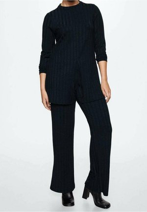 Person iført en sort ribstrikket tunika med langærmer og frontslids, matchende bukser med vide ben og sorte ankelstøvler med hæl på hvid baggrund.