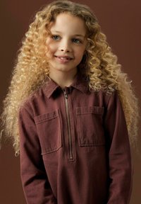 Chemise zippée bordeaux avec deux poches avant et un col, fabriquée en tissu texturé ; le mannequin a de longs cheveux blonds bouclés.
