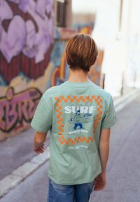 Personne aux cheveux courts portant un t-shirt vert "Surf Club" avec une bordure à carreaux orange, marchant dans une ruelle urbaine aux murs tagués.
