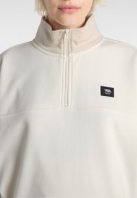 Polaire zippée beige avec un col haut et un petit logo Vans noir sur la poitrine, présentant un tissu lisse et texturé.