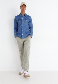 Camicia di jeans azzurra chiara con bottoni frontali e due tasche sul petto, abbinata a pantaloni beige chiaro e sneakers bianche con dettagli rossi.