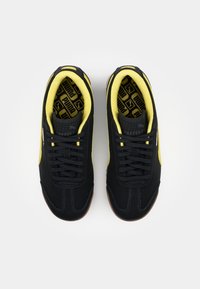 Baskets Puma noires et jaunes avec des lacets noirs, une doublure intérieure jaune et des semelles intérieures marquées, vues d'au-dessus sur un fond blanc.