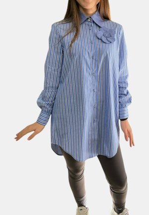 Donna che indossa una camicia lunga a righe verticali blu e bianche con una spilla a forma di fiore di stoffa, abbinata a leggings scuri e sneakers bianche.