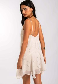 Vestido beige claro con tirantes finos, que presenta un diseño en capas y acentos metálicos. La tela transparente y texturizada añade dimensión a la prenda.