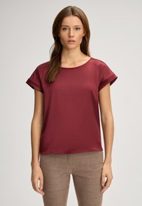 Bordeauxfarbiges kurzärmeliges Oberteil mit rundem Ausschnitt und glatter Textur, kombiniert mit karierten braunen Hosen mit roten Akzenten.