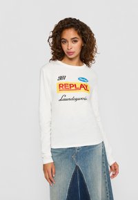 Vrouw met krullend haar, gekleed in een witte blouse met lange mouwen met de tekst "REPLAY" en een twee-kleurige denim rok, staand tegen een effen achtergrond.